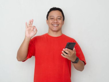 Kırmızı tişörtlü genç Asyalı adam beyaz arka plan üzerinde telefonunu tutarken iyi bir el hareketi yapıyor. Görüntü pozitifliği ve onayı tasvir ediyor, tatmin kavramları için mükemmel.