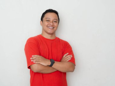 Kırmızı tişörtlü gülümseyen Asyalı genç adam, kollarını kavuşturmuş beyaz arka planda duruyor..