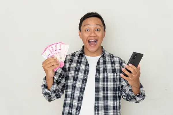 Genç Asyalı adam, elinde cep telefonu ekranı ve Rupiah parası tutarken heyecanlı ve mutlu. Boşluğu kopyala