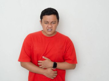 Young Asian man in red shirt clutches his stomach, showing discomfort or pain. Man suffering from stomach ache over white background. clipart