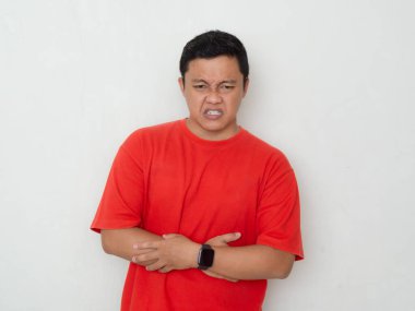 Young Asian man in red shirt clutches his stomach, showing discomfort or pain. Man suffering from stomach ache over white background. clipart