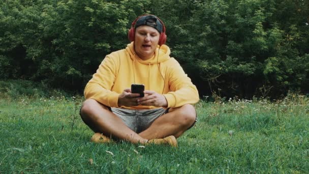 一个戴耳机的年轻人坐在大自然中 一边听音乐一边哼着他最喜欢的歌 戴双耳耳机的男人 — 图库视频影像