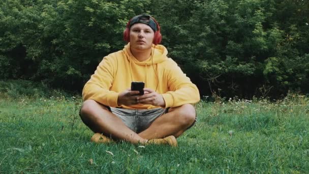 忧郁的年轻人听着大自然的音乐 跳着舞动着他的身体 坐在草地上 头上戴红色耳机的青少年 — 图库视频影像