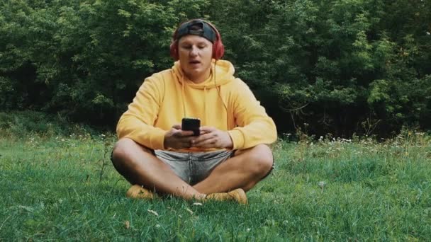 快乐的年轻人在大自然中听音乐 头上戴耳机的青少年坐在草地上 一个人一边听音乐一边哼着歌 — 图库视频影像