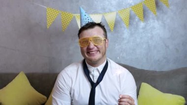 Elinde maytap, şenlik şapkası, gömleği ve garip kravatıyla doğum gününü kutlayan komik bir genç adam. Neşeli, komik, gülümseyen, mutlu genç adam tatili neşeli bir şekilde kutluyor.
