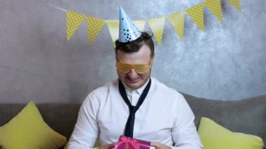 Kafasında şenlik şapkası olan, esnek kravatlı, neşeli bir ofiste çok güzel gülümseyen ve doğum günü hediyesini açan tuhaf bir adam. Komik hediyeli pembe bir kutu. 