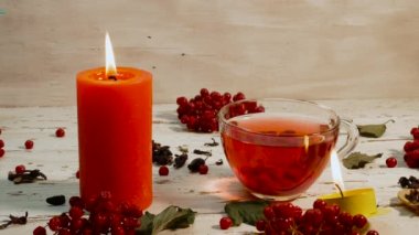 Beyaz ahşap bir masada kırmızı çay ve yanan mumlarla dolu bir fincan. Kırsal panolardaki kırmızı viburnum üzümünün doğal çayı. Naturopati iyileşme aşısı. Kırmızı böğürtlenlerden yapılan doğal antioksidan.