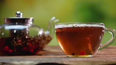 Yavaş çekimde, şeffaf cam bardakta çaydanlık ile meyveli meyveli çilek. Yeşil Bokeh arka planında doğal çay. Kırmızı viburnum ve siyah yaban mersinli çay
