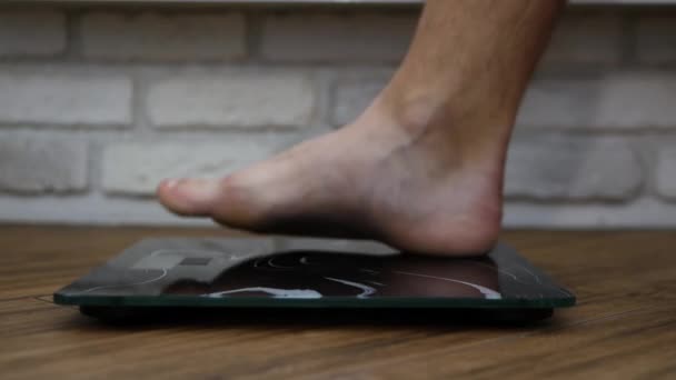 裸足の男の足は 白いレンガの壁を背景に床の上に立つ 人は体重計で量られます 減量の概念 体重計の男の体 — ストック動画