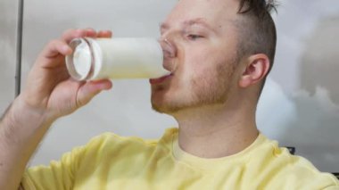 Genç bir adam iştah açıcı bir şekilde taze süt ya da şeffaf bir bardaktan milkshake içer. Kefir ya da süt ürünü içen bir adamın portresi. Vücuttaki kalsiyum kaynağını bir ürünle doldurmak.