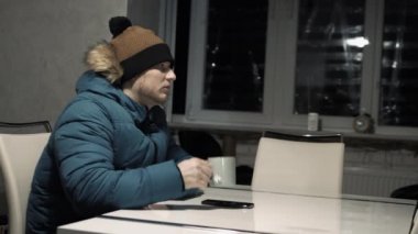 Genç bir adam evde ısınırken çay içiyor. Adam evde donup kalıyor. Ceketli ve şapkalı bir adam evde kendini ısıtıyor.