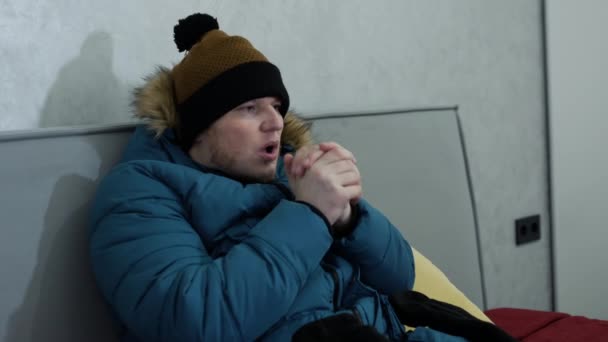 一个年轻人在家里因寒冷而冻僵了 一个男人坐在沙发上的时候戴上了温暖的手套 一个穿着夹克和帽子的男人在家里沙发上 在世界天然气危机背景下取暖的问题 — 图库视频影像