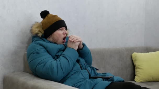一个人在家里因寒冷 煤气危机而冻僵了 一个戴冬季手套的男人正坐在沙发上用智能手机打字 一个穿着夹克和帽子的男人在家里 在天然气危机背景下取暖的问题 — 图库视频影像