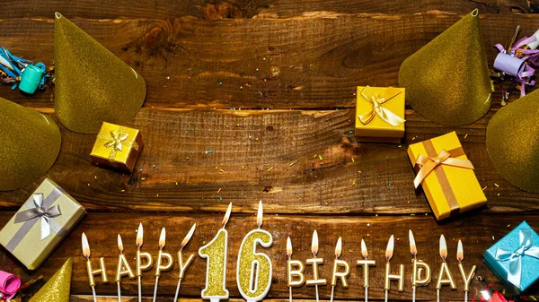 Top view of the golden letters of the candle with the number happy birthday, the background of the gift boxes with candles happy birthday on the background of brown boards. Copy spacePostcard Happy birthday 16.