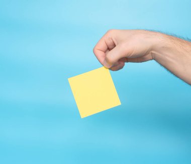 A hand holds a yellow blank letter sticker for the inscription of your text. Message on sticker copy space. Mini banner for text in a person's hand. On a pastel blue background