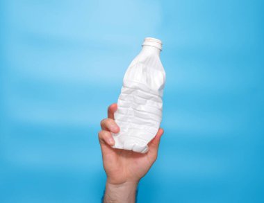 Plastic trash concept. A twisted squeezed used plastic white bottle in a man's hand on a blue background.