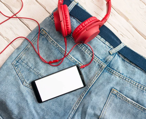 The concept of listening to music in a smartphone, on jeans the display of a smartphone is white with headphones from above view. Red overhead musical headphones.