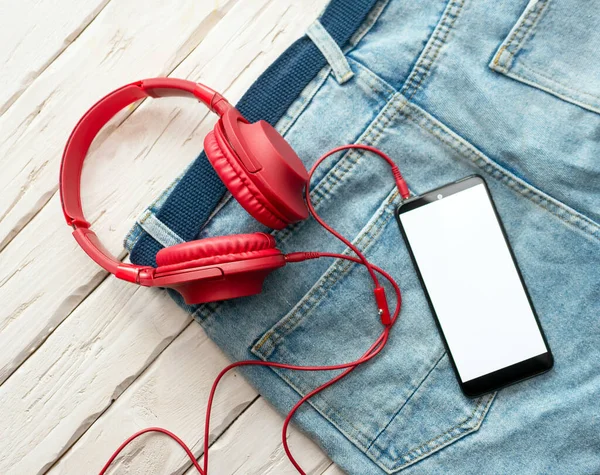 The concept of listening to music in a smartphone, on jeans smartphone display white copy space for text with headphones top view. Red overhead musical headphones.