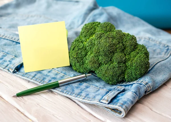 A bunch of Broccoli with a notepad or sticker yellow blank for text in a jeans pocket. Green tree on denim. Notes for inscription