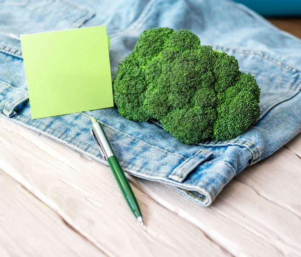 A bunch of Broccoli with a notepad or sticker yellow blank for text in a jeans pocket. Green tree on denim. Notes for the inscription about the vitamins of cabbage