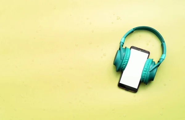 Mint on-ear music headphones with a smartphone with a white display. Headphones with a gadget on a yellow background in water drops. top view