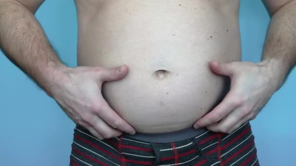 用手触摸厚厚的腹部脂肪层 一个肥胖的年轻人正试着穿牛仔裤裤 一个大肚皮的男人肥胖的人减肥的概念 蓝色背景的演播室视频 — 图库视频影像