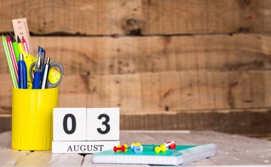 Ağustosta 3 numaralı takvim. Ahşap bir arka planda bir çantada kırtasiye kalemleri ve kalemler. Yer defterini kalem ve takvimle kopyala. Metin için planlayıcı yer.