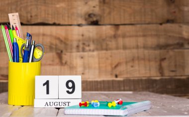 Ağustos takviminde 19 numara var. Ahşap bir arka planda bir çantada kırtasiye kalemleri ve kalemler. Yer defterini kalem ve takvimle kopyala. Metin için planlayıcı yer.