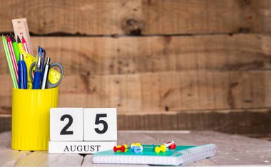 Ağustos takviminde 25 numara var. Ahşap bir arka planda bir çantada kırtasiye kalemleri ve kalemler. Yer defterini kalem ve takvimle kopyala. Metin için planlayıcı yer.