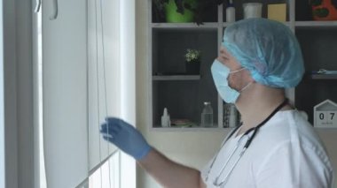 Doktor pencereden dışarı bakar. Steteskoplu, tıbbi maskeli ve steril şapkalı bir çocuk doktoru. Ofisteki doktor ofisteki pencerenin perdelerini sökmüş.