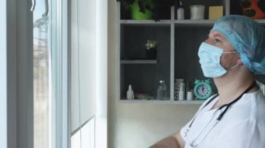 Doktor pencereden dışarı bakar. Tıbbi maske takmış steteskoplu ve steril şapkalı bir çocuk doktoru.