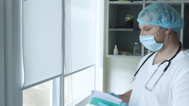 彼の手にノートがある医者 医療用マスクと滅菌キャップの聴診器を備えた小児科医 オフィスの医者は患者への治療のための処方箋を書いています — ストック動画