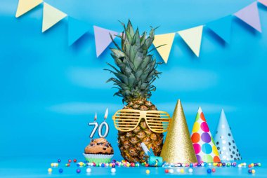Güneş gözlüklü ananas karakterli yaratıcı arka plan fotokopi alanı. Doğum günün kutlu olsun muffin ile mi yoksa mum numarası 70 olan pastayla mı? Mavi arka planda yıldönümü süslemeleri.