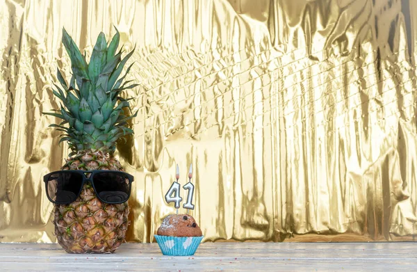 Cheerful pineapple in glasses festive happy birthday character with number  41. Beautiful background for congratulations Koiya space on a golden shiny background. Anniversary congratulations with the number of candles burning with a muffin.