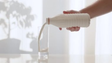 Plastik şişeden bir bardağa süt dök. Arka plan sütü beyaz kadife perdelerin arka planına dökülüyor. Mutfak masasındaki şeffaf bardakta taze süt ürünü..