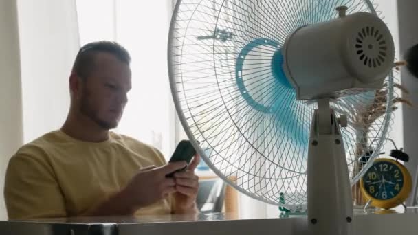 スマートフォンを持ったハンサムな若者 アパートは暑い夏のファンによって冷却されます アパートのエアダクトファンブレード ウインドブレード — ストック動画