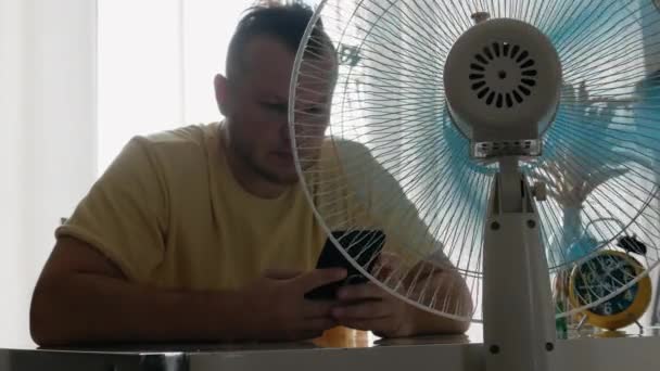 电风扇使空气变冷 这个人很热 风扇是公寓里的通风管 男人手里拿着智能手机冷静下来 — 图库视频影像