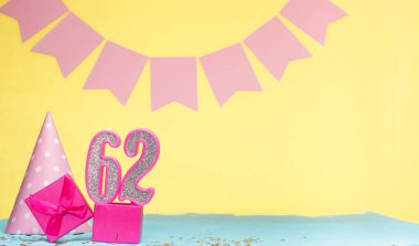 62 yaşında bir kızın doğum tarihi. Uzayı kopyala Sarı arkaplanlı pembe gözlüklü bir doğum günü. Üzerinde numaralandırılmış mumlar ve hediye kutusu olan dekorasyonlar. Bir kadın için yıldönümü kartı