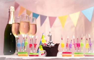 Şampanya ve bardaklarla doğum günü. Doğum günü mumu, 1 numara. Yıldönümü kartı ve çelenkler yer açıyor. Neşeli arkaplan.