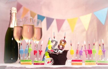Şampanya ve bardaklarla doğum günü. 28 numarayla doğum günü mumu. Yıldönümü kartı ve çelenkler yer açıyor. Neşeli arkaplan.