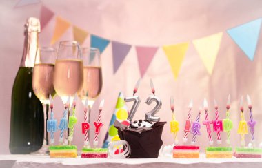 Şampanya ve bardaklarla doğum günü. 72 numarayla doğum günü mumu. Yıldönümü kartı ve çelenkler yer açıyor. Neşeli arkaplan.