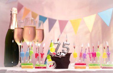 Şampanya ve bardaklarla doğum günü. 75 numarayla doğum günü mumu. Yıldönümü kartı ve çelenkler yer açıyor. Neşeli arkaplan.
