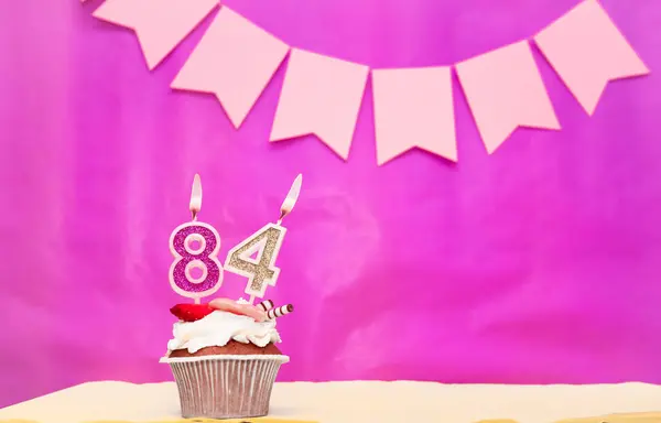 Background date of birth with number  84. Pink background with a cake and burning candles, save space, happy birthday anniversary for a girl. Holiday pudding muffin.