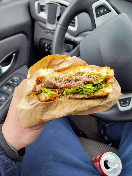 A man eats fast food in the car, a burger in the hand of the men in the car. Snack food in the vehicle. Eating while driving