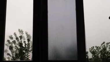 Pencerenin dışında yağmur, arka planda yeşil ağaçların arasında yaz yağmuru. Pencere camında yağmur damlaları