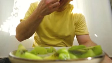 Genç adam bir tabaktan kesilmiş parçalarla kivi meyvesi yiyor, ağır çekimde.