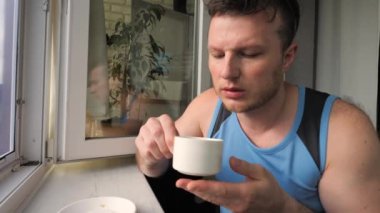 Genç bir adam pencerenin kenarında oturuyor ve beyaz bir fincandan koyu kahve içiyor..