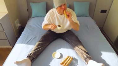 Yakışıklı ve şık bir genç adam yatakta kahvaltı yapıyor. Yatak odasında, yatakta sade kahve içmek. Yoğunlaştırılmış sütle waffle böreği ye. Yavaş çekim.