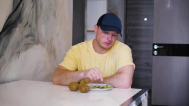Genç adam mutfakta otururken ağır çekimde kivi meyvesi yiyor.