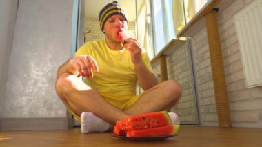 Genç bir adam bir dairede olgun bir karpuz yiyor. Elinde karpuz olan sarı elbiseli bir adam. Yavaş çekim